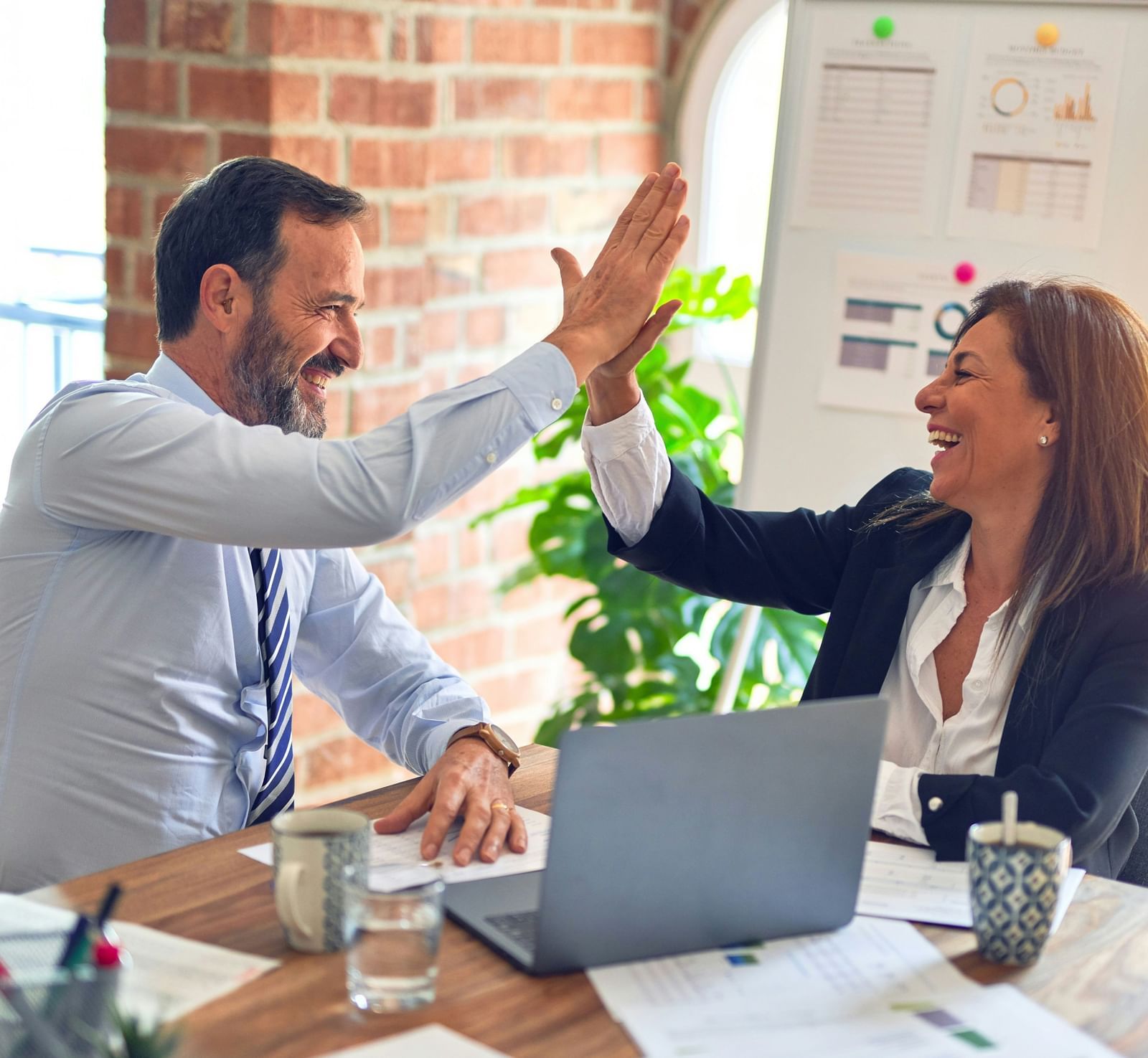 Two colleagues in a bright, modern office space sharing a high-five over a desk cluttered with papers, notebooks, and a laptop.